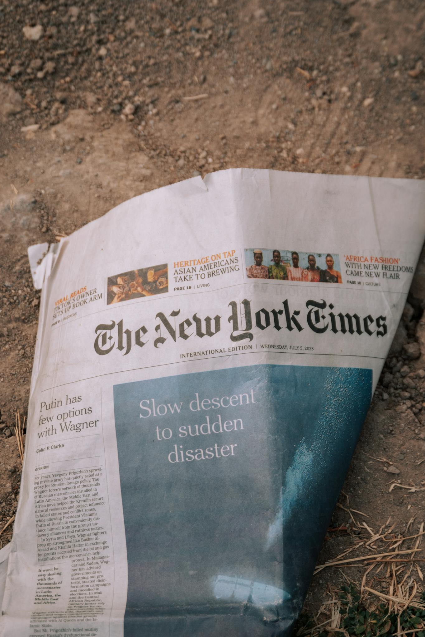 A wrinkled The New York Times newspaper lies on a dirt surface outdoors.